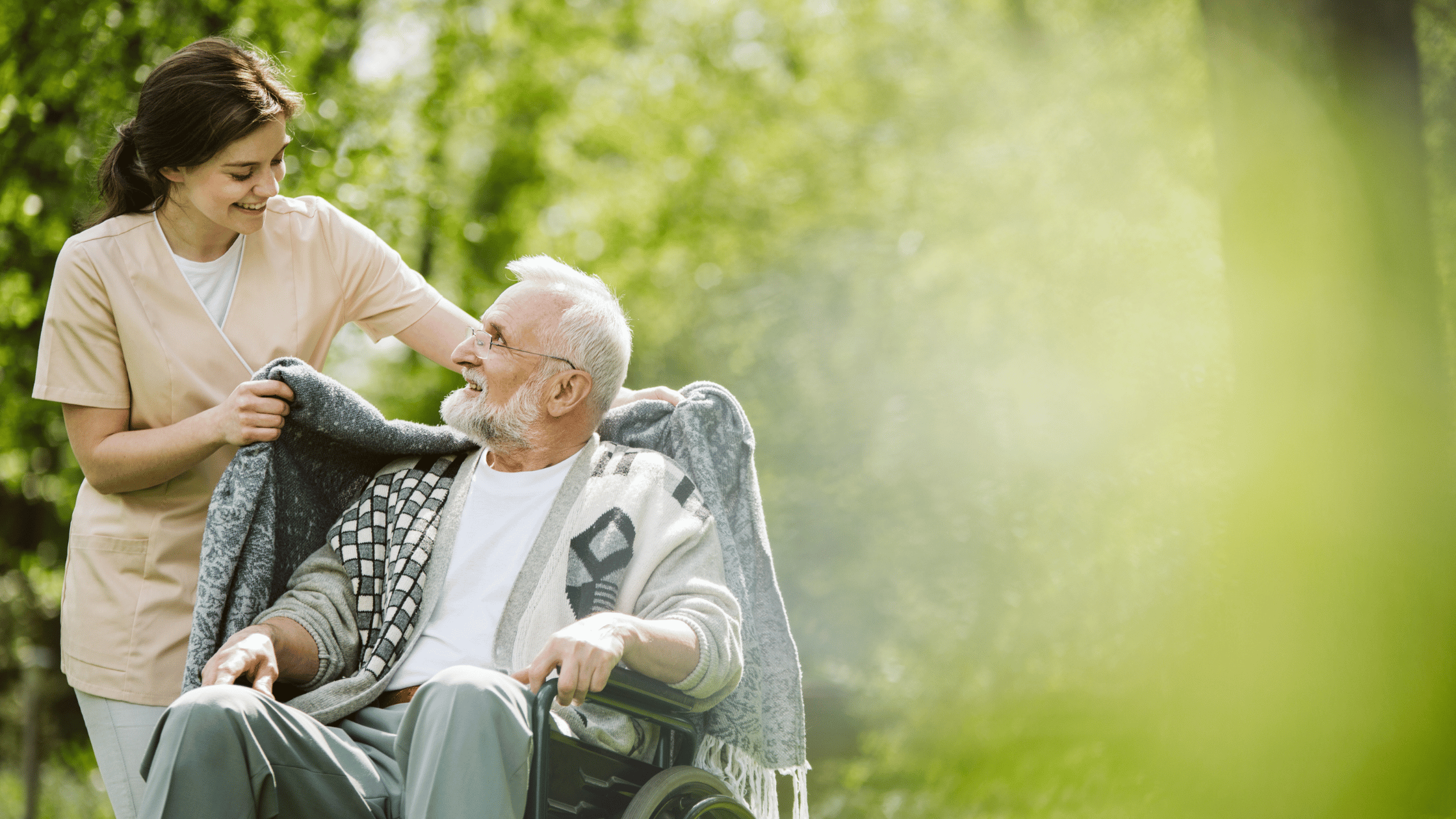 An elderly man in a wheelchair being pushed by a young aged care worker.